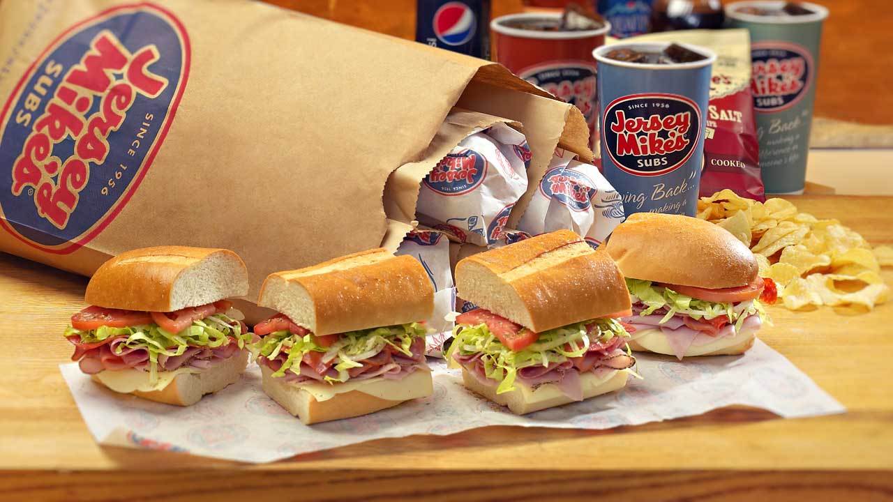 Fresh Jersey Mike's subs with a branded to-go bag, cups, and chips